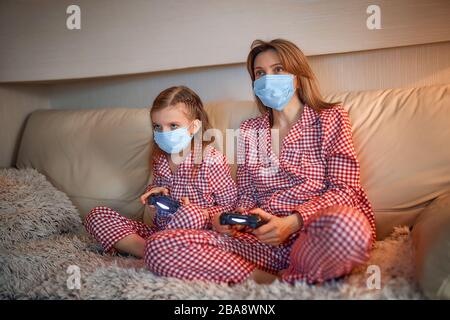 Femme et jeune fille portant un pyjama et des masques de protection médicale assis sur un canapé dans le salon avec des contrôleurs de jeu vidéo à domicile isolation auto quarantaine, covid-19. Banque D'Images