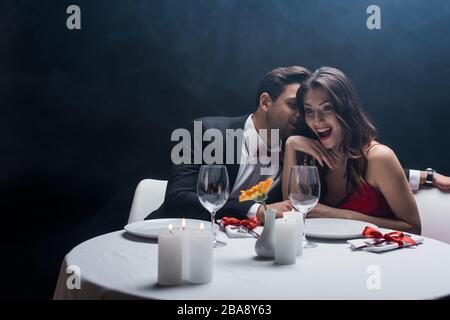 Un homme en costume chuchoté à une femme choquée à table servie sur fond noir avec de la fumée Banque D'Images