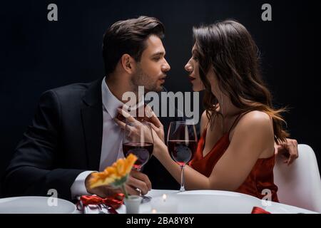 Vue latérale de la belle femme ajustant noeud papillon de beau homme pendant le dîner romantique isolé sur noir Banque D'Images