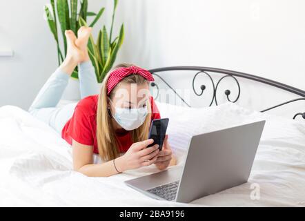 une jeune femme malade dans un masque médical et un t-shirt rouge et un pantalon confortable se trouvent sur un lit blanc avec un ordinateur portable et un téléphone. elle travaille de la maison parce que Banque D'Images