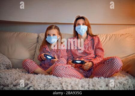 Femme et jeune fille portant un pyjama et des masques de protection médicale assis sur un canapé dans le salon avec des contrôleurs de jeu vidéo à domicile isolation auto quarantaine, covid-19. Banque D'Images