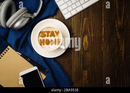 Lieu de travail à la maison. Tasse de café avec texte RESTER MAISON sur table en bois avec tissu bleu. Latte art doit faire l'apparition d'une éclosion de quarantaine de covid-19. Banque D'Images