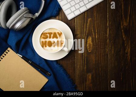 Lieu de travail à la maison. Tasse de café avec texte RESTER MAISON sur table en bois avec tissu bleu. Latte art doit faire l'apparition d'une éclosion de quarantaine de covid-19. Banque D'Images