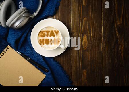 Lieu de travail à la maison. Tasse de café avec texte RESTER MAISON sur table en bois avec tissu bleu. Latte art doit faire l'apparition d'une éclosion de quarantaine de covid-19. Banque D'Images