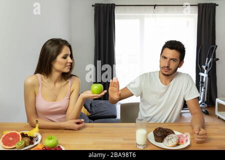 Un homme mène un mauvais style de vie, il mange des beignets avec givrage pour le petit déjeuner. La fille lui offre d'essayer des fruits frais, mais il refuse. La confrontation de Banque D'Images