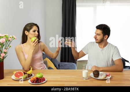 Un homme mène un mauvais style de vie, il mange des aliments sucrés et riches en calories pour le petit déjeuner. Il offre à la fille d'essayer un beignet de chocolat, mais elle refuse parce que Banque D'Images