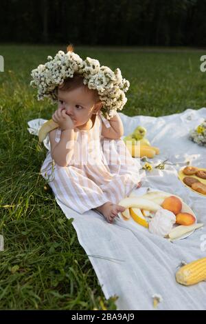 Petite fille dans une robe rayée sur un pique-nique dans un parc de la ville. Elle mange de la baguette fraîche et des fruits, boit du jus naturel. L'harmonie de l'homme et de la nature. Banque D'Images