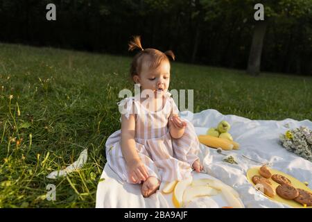 Petite fille dans une robe rayée sur un pique-nique dans un parc de la ville. Elle mange de la baguette fraîche et des fruits, boit du jus naturel. L'harmonie de l'homme et de la nature. Banque D'Images