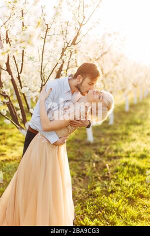 Beau couple, gai et romantique amoureux se posant dans le jardin fleuri. Bel homme embrassant le cou de sa jolie femme dans une robe élégante avec de grandes Banque D'Images