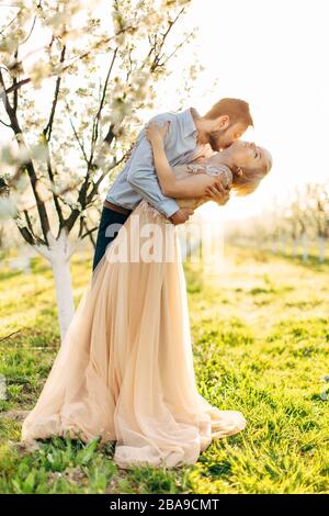 Beau couple, gai et romantique amoureux se posant dans le jardin fleuri. Bel homme embrassant le cou de sa jolie femme dans une robe élégante avec de grandes Banque D'Images