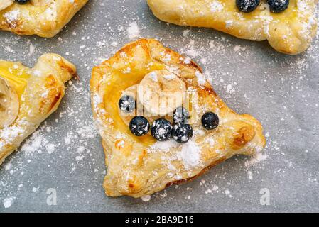 Biscuits à base de pâte feuilletée avec pouding, banane et bleuets saupoudrés de sucre en poudre couché sur du papier de cuisson. Banque D'Images