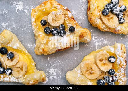 Biscuits à base de pâte feuilletée avec pouding, banane et bleuets saupoudrés de sucre en poudre couché sur du papier de cuisson. Banque D'Images