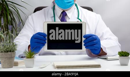 médecin homme assis à un bureau dans un manteau médical blanc et tenant une tablette électronique avec un écran noir vierge, portant des gants médicaux bleus, en ligne Banque D'Images