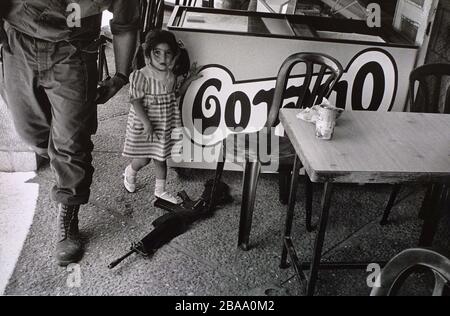 Une jeune fille de colon pointant vers un fusil appartenant à un soldat de la Force de défense d'Israël (FDI) dans un café de la colonie de Kiryat Arba, en Cisjordanie, près d'Hébron. En 1994, un colon nommé Baruch Goldstein a tué 29 Palestiniens dans la ville voisine d'Hébron. Israël a occupé la Cisjordanie, Gaza et le plateau du Golan au lendemain de la guerre de six jours de 1967. Banque D'Images