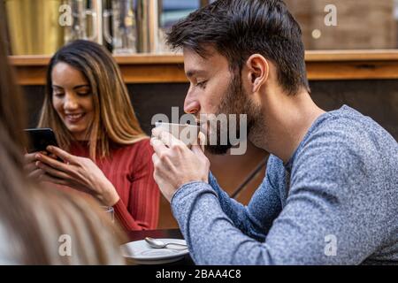 Groupe d'amis au café. Un jeune adulte barbu du cappuccino à partir d'un mug blanc. D'autres amis souriant en regardant leurs smartphones. Hors-faire Banque D'Images