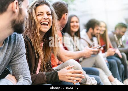 Groupe de jeunes branchés discutant ensemble assis sur un banc à l'extérieur. Les étudiants s'amusent ensemble. Concentrez-vous sur une jeune fille blonde souriante avec un protocole d'entente ouvert Banque D'Images