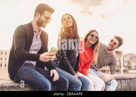 Groupe de jeunes branchés discutant ensemble assis sur un banc à l'extérieur. Les étudiants s'amusent ensemble. Concentrez-vous sur une fille brunette souriant avec o Banque D'Images