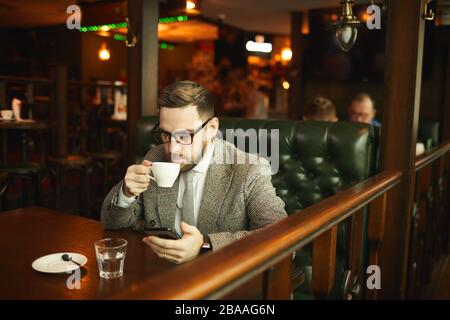 Jeune homme d'affaires dans des lunettes et en costume boire du café et utiliser son téléphone mobile tout en étant assis à la table dans le café Banque D'Images