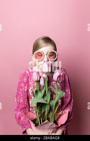 Blonde Tween fille dans des lunettes de soleil sent des tulipes roses sur fond rose Banque D'Images