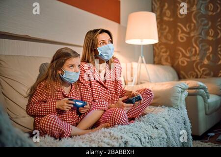 Femme et jeune fille portant un pyjama et des masques de protection médicale assis sur un canapé dans le salon avec des contrôleurs de jeu vidéo à domicile isolation auto quarantaine, covid-19. Banque D'Images