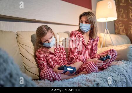 Femme et jeune fille portant un pyjama et des masques de protection médicale assis sur un canapé dans le salon avec des contrôleurs de jeu vidéo à domicile isolation auto quarantaine, covid-19. Banque D'Images