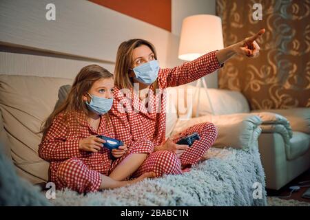 Femme et jeune fille portant un pyjama et des masques de protection médicale assis sur un canapé dans le salon avec des contrôleurs de jeu vidéo à domicile isolation auto quarantaine, covid-19. Banque D'Images