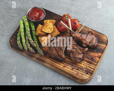 Côtelettes d'agneau grillées sur un panneau en bois avec asperges, sauce rouge, tomates Banque D'Images