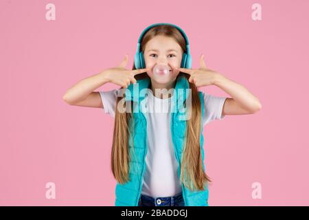 Une petite fille avec casque explose la gomme à mâcher rose sur un fond rose. Banque D'Images