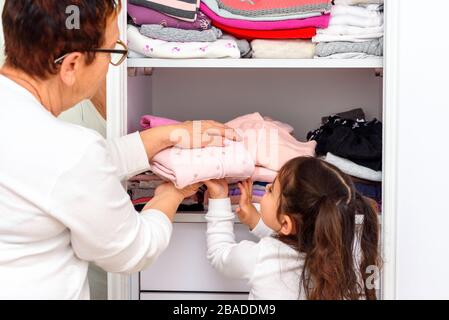 Femme senior avec sa petite-fille qui stocke des vêtements dans la garde-robe. Les enfants aident grand-mère en pliant la lessive. Jeune fille sur l'étude à la maison en raison de l'éclosion de Covid 19. Banque D'Images