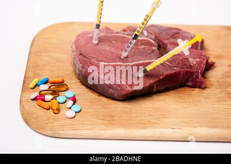 Seringues dans la viande crue et les pilules hormonales sur le tableau de découpe isolé sur blanc Banque D'Images