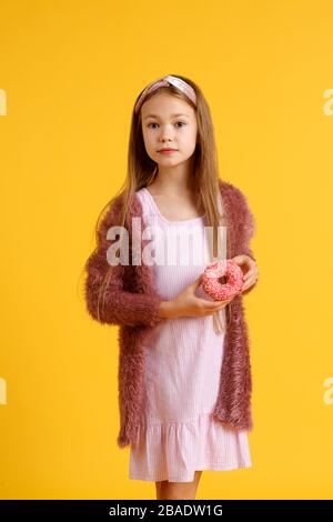 Une jeune fille blonde folle et gaie dans un chapeau d'anniversaire souriant, s'amuser et regarder par deux beignets rouges sur ses yeux. Bonbons. Fond de studio jaune. Banque D'Images