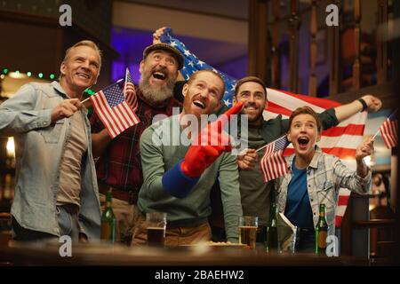 Groupe de fans américains debout à la table avec boissons alcoolisées et regarder le match de l'équipe de football dans le bar sportif Banque D'Images