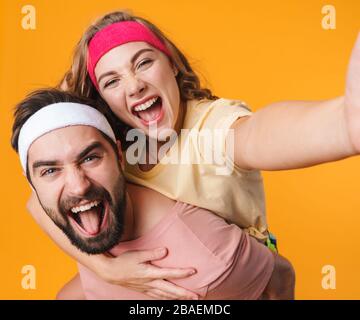 Portrait de jeune couple sportif heureux portant des bandeaux souriants tout en faisant une balade en piggyback isolée sur fond jaune Banque D'Images