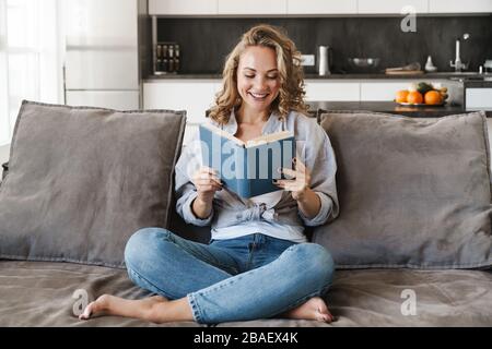 Jeune blonde gaie une femme qui a lu un livre tout en étant assise sur un canapé à la maison Banque D'Images