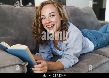Jeune blonde gaie une femme qui lit un livre tout en se posant sur un canapé à la maison Banque D'Images