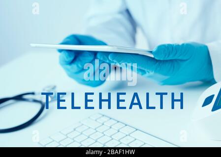 le texte télésanté et un médecin, portant un manteau blanc et des gants chirurgicaux bleus, à l'aide d'une tablette numérique assise à un bureau Banque D'Images