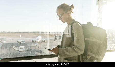 Jeune femme se tenant près des portes de l'aéroport, tenant le téléphone portable dans ses mains, portant un sac à dos de voyage et marchant jusqu'au salon. Voyageur féminin Banque D'Images