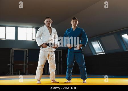 Deux jeunes combattants caucasiens judo en kimono blanc et bleu avec ceintures noires posant confiance dans la salle de gym, forte et saine. Pratiquer des habiletés de combat en arts martiaux. Surmonter, atteindre l'objectif. Banque D'Images
