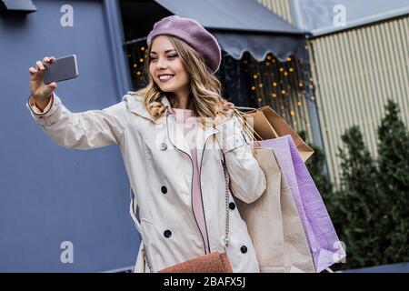 Partage de photos avec des amis. Photo horizontale d'une belle femme blonde en béret prenant selfie sur smartphone et souriant tout en se tenant avec des magasins b Banque D'Images