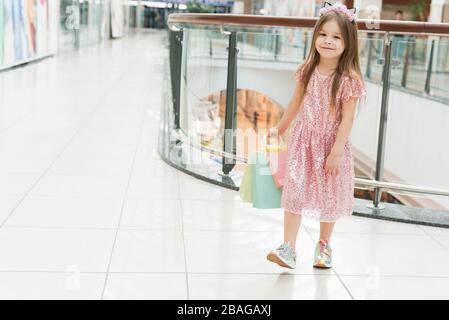 Portrait d'une petite fille heureuse dans le centre commercial. Une fille souriante riant dans une robe rose avec des sacs multicolores dans ses mains marche autour du centre commercial, en regardant les fenêtres de magasin. Banque D'Images