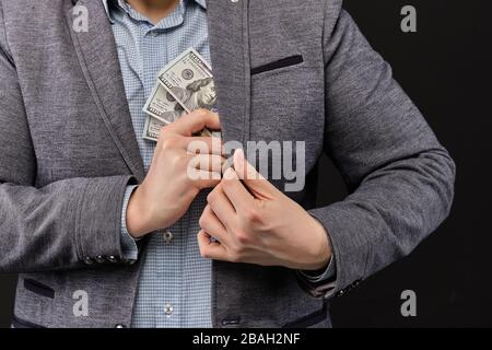 Un homme dans une veste met des dollars dans sa poche sur un fond noir. Gros plan. Banque D'Images
