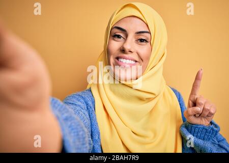 Jeune femme arabe brunette portant le hijab islamique faire selfie par caméra surpris par une idée ou une question pointant le doigt avec le visage heureux, nu Banque D'Images