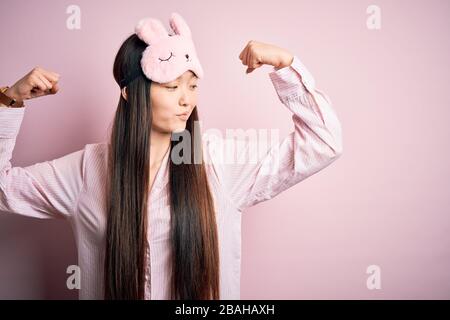 Jeune femme asiatique portant un masque de pyjama et de sommeil sur fond rose isolé montrant les muscles des bras souriant fier. Concept de forme physique. Banque D'Images