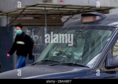 Bucarest, Roumanie - 17 mars 2020: Un taxi britannique poussiéreux, un foyer sélectif, et un jeune homme passant et porter un masque de protection contre le coronavirus Banque D'Images