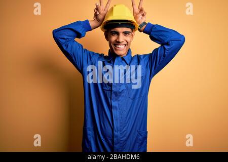 Jeune homme de travail africain américain portant un uniforme bleu et un casque de sécurité posant drôle et folle avec les doigts sur la tête comme des oreilles de lapin, souriant Banque D'Images