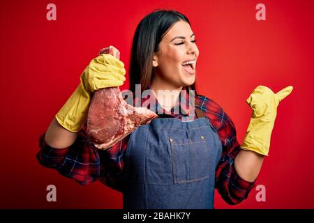 Jeune boucher femme tenant de la viande de boeuf fraîche brute piquer sur fond rouge pointant et montrant avec le pouce jusqu'au côté avec le visage heureux souriant Banque D'Images