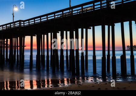 Aperçu de Pier au coucher du soleil à Ventura, Californie. Roches et sable en premier plan, l'eau s'est replacée en marée basse. Lampes sur la jetée ; ciel crépuscule coloré à l'arrière Banque D'Images