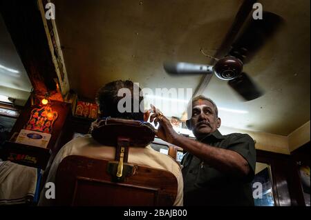 un barbier qui tend à un client sous un ventilateur dans son magasin , Madurai, Inde Banque D'Images