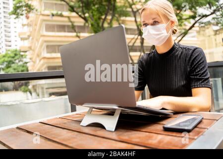 Blonde femme avec masque de protection sur son visage travaillant à distance avec son ordinateur portable dans une table en bois avec un smartphone dans sa terrasse extérieure. Covid-19 Banque D'Images