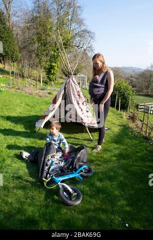 Mère avec porte-bébé en harnais et enfants de garçon avec tipi jouant dans un jardin de campagne dehors au printemps 2020 Pays de Galles UK KATHY DEWITT Banque D'Images